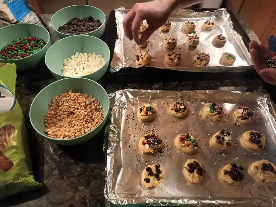 A person is decorating raw cookie dough balls on two foil-lined baking sheets with various toppings. Four light green bowls…