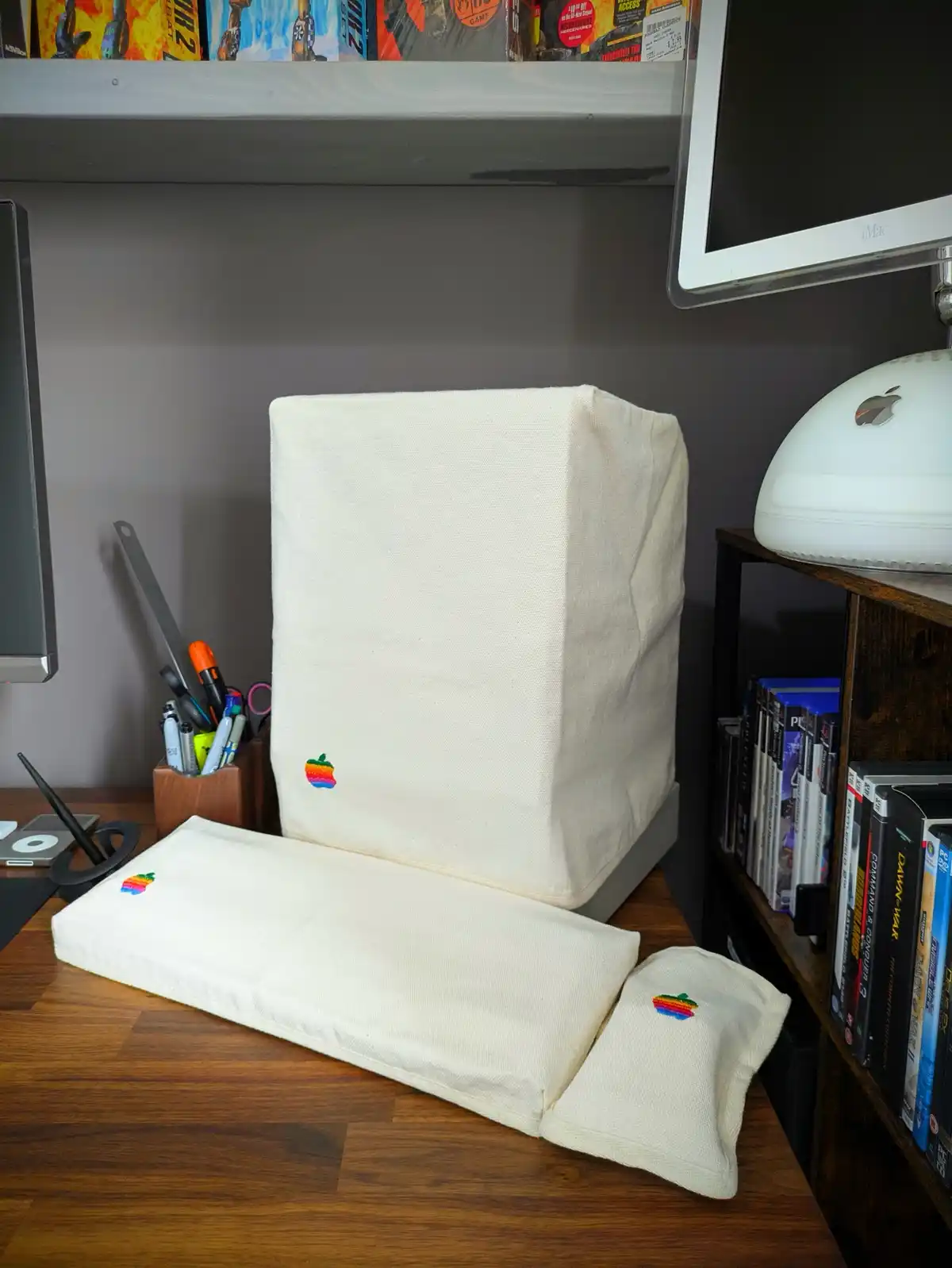 A desk setup features a vintage Apple computer tower and keyboard covered in off-white fabric with rainbow Apple logos…
