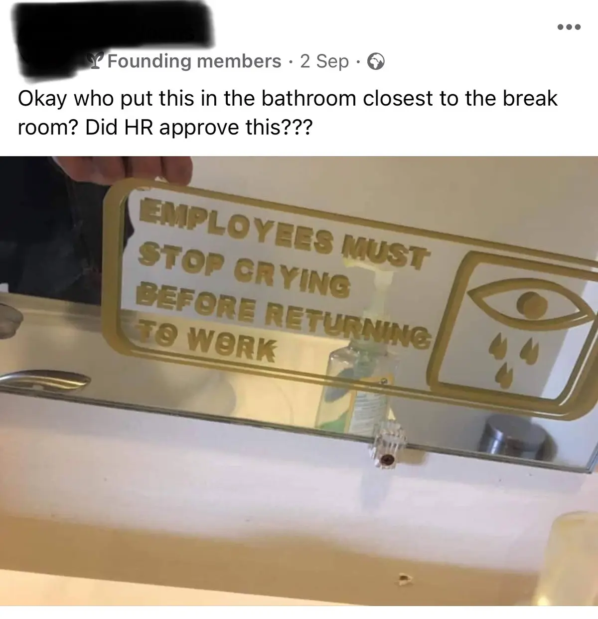 A person holds up a gold-framed sign in a bathroom that reads 'EMPLOYEES MUST STOP CRYING BEFORE RETURNING TO WORK' with an…