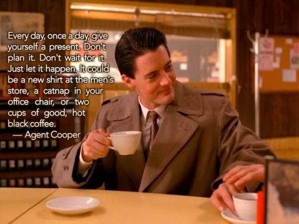 A still from the TV show Twin Peaks shows Agent Cooper sitting at a diner table, holding a coffee cup, with a calm, slightly…