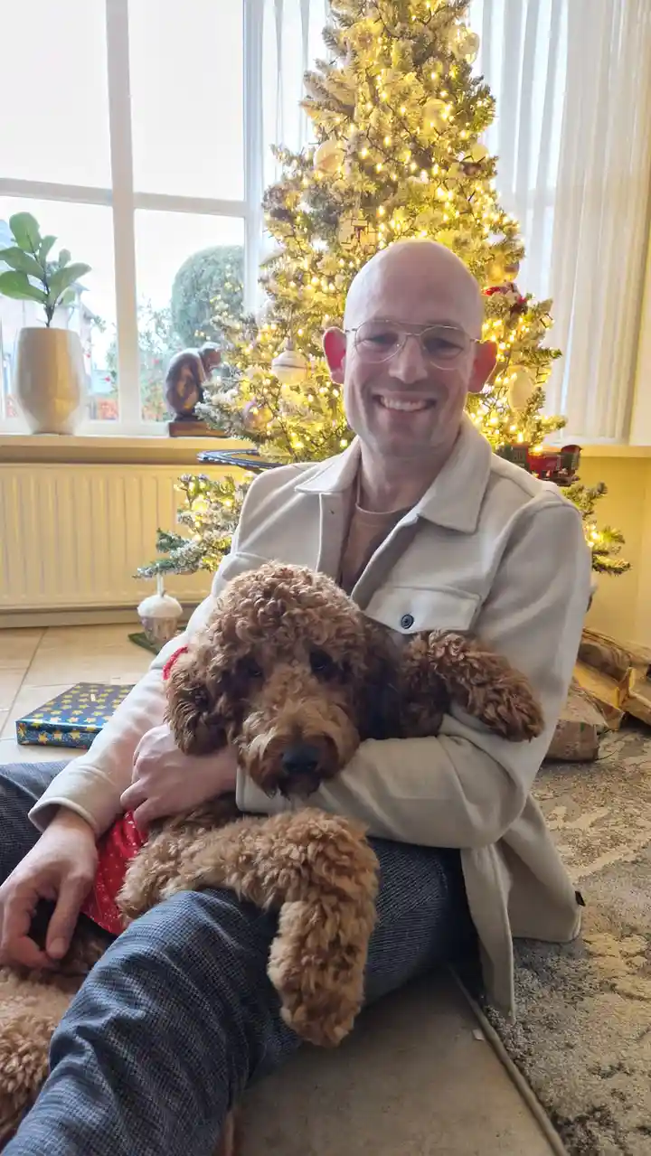 A bald man with glasses sits on the floor in front of a lit Christmas tree, smiling while holding a brown curly-haired dog…