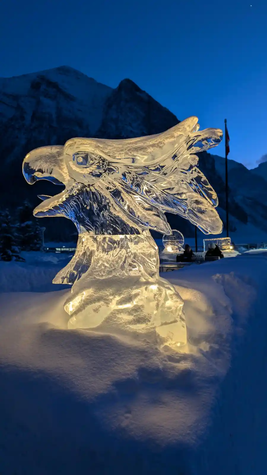 A large, intricately carved ice sculpture of a bird's head, possibly an eagle or hawk, is illuminated from within and set in…