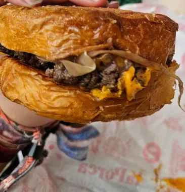 A close-up photo shows a hand holding a large, messy burger with a toasted bun, melted cheese, beef patty, and onions. The…