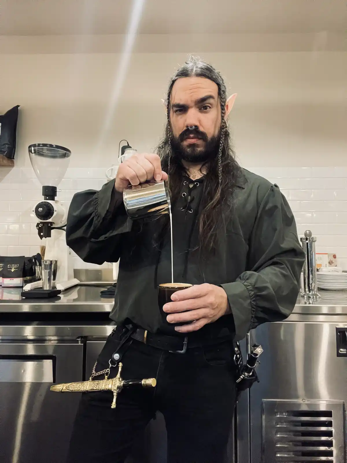A man with long dark hair, pointed ears, and a nose ring pours milk into a coffee cup in a modern cafe setting. He wears a…
