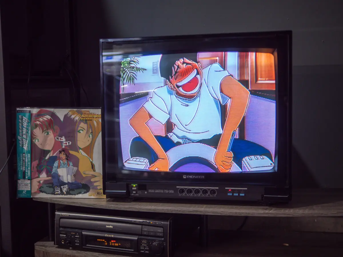 A vintage Pioneer CRT television displays an anime scene of a character laughing maniacally while driving, next to a…