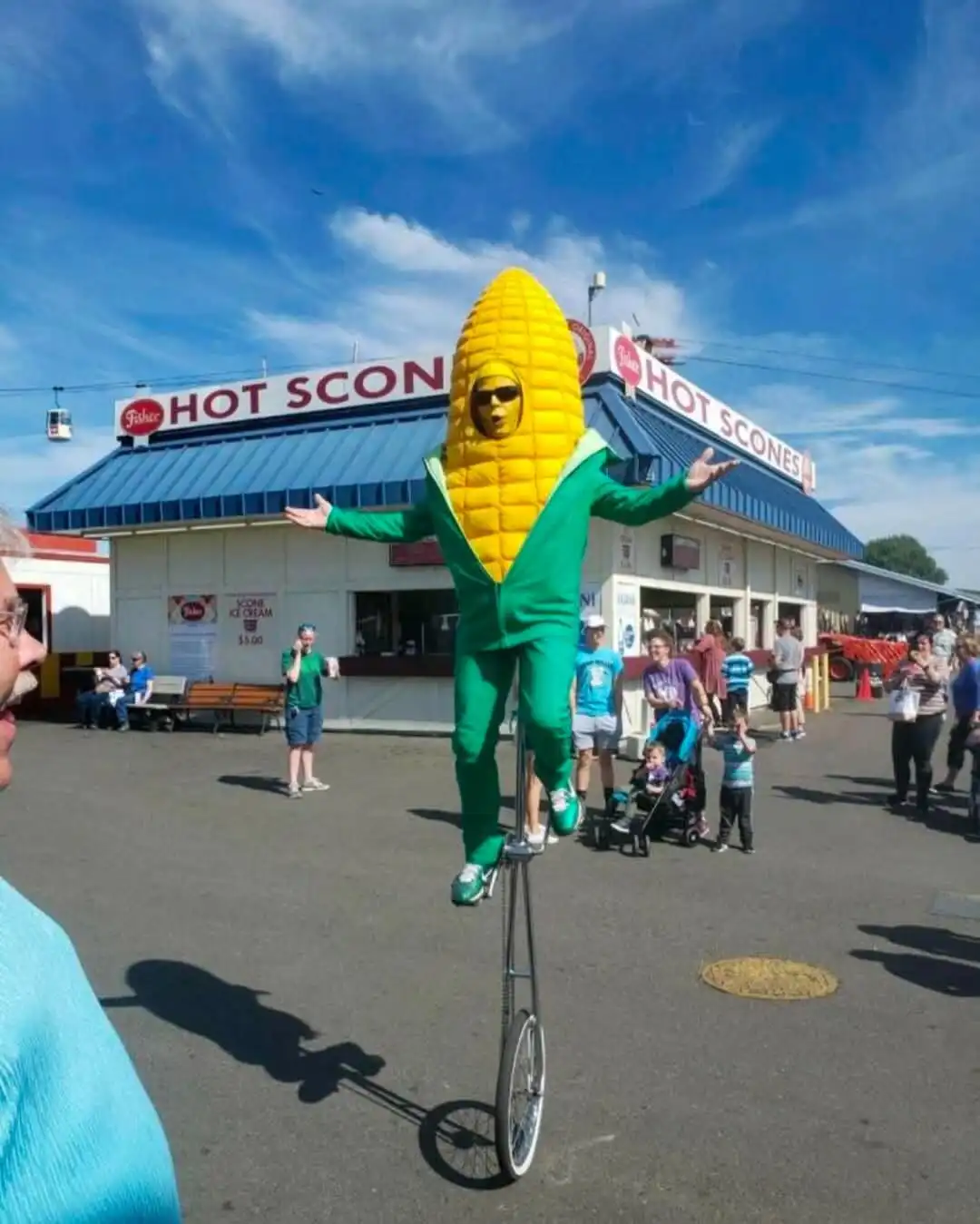 A person in a large, yellow corn-on-the-cob costume with a green jumpsuit and sunglasses is balancing on a unicycle in front…
