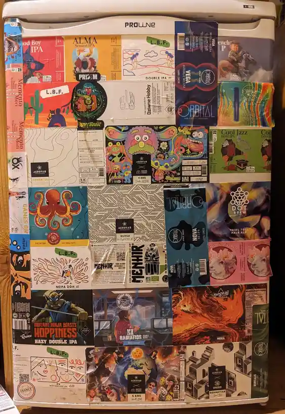 A white Proline mini-fridge is completely covered in colorful, overlapping beer labels from various craft breweries…