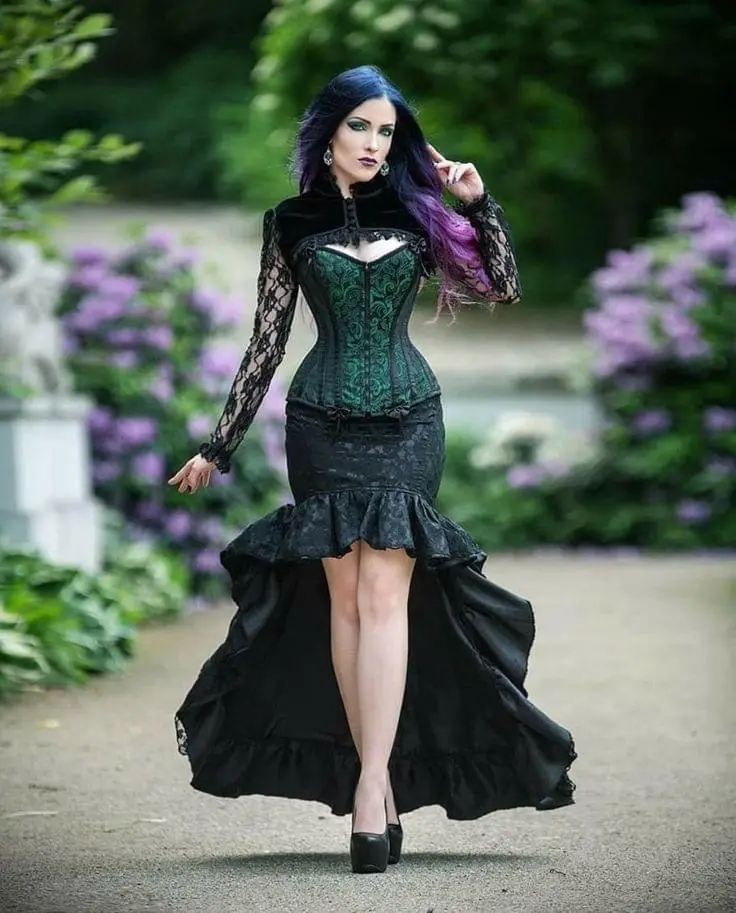 A woman with dark blue and purple hair, wearing a green corset and black lace dress with a high-low skirt, poses outdoors on…