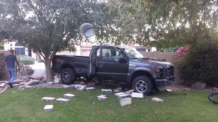 A black pickup truck is stuck in a yard, surrounded by broken concrete pavers, with a man walking away from the scene. A…