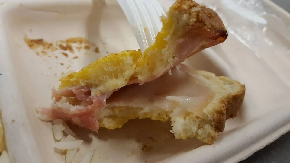A close-up photo shows a fork holding a partially eaten sandwich made with bread, ham, and melted cheese, resting on a white…