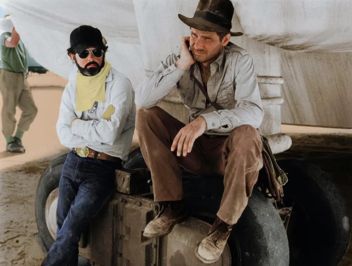 Two men sit on a vehicle in a desert setting; one wears a fedora and holds a phone to his ear with a skeptical expression…