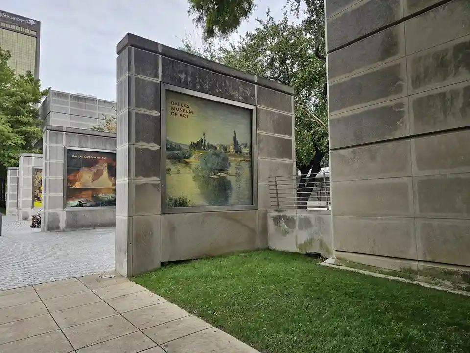 An outdoor view of the Dallas Museum of Art, showing stone walls with framed paintings displayed on them, including one of a…