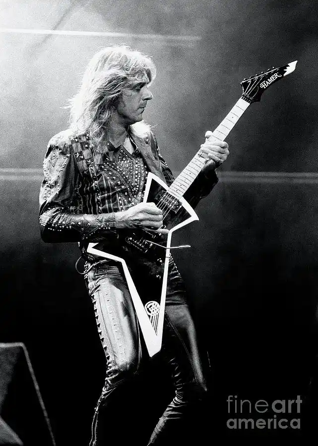 A black-and-white photo of a man with long hair playing a distinctive star-shaped electric guitar on stage. The guitar has a…