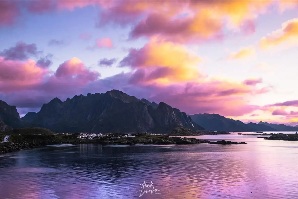 A scenic photograph of a fjord at sunset, featuring dramatic purple and orange clouds reflecting on calm water, with rugged…