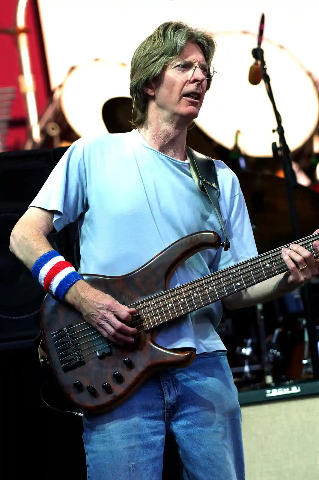 A man with glasses and shoulder-length hair plays a bass guitar on stage while wearing a light blue t-shirt and jeans. He…