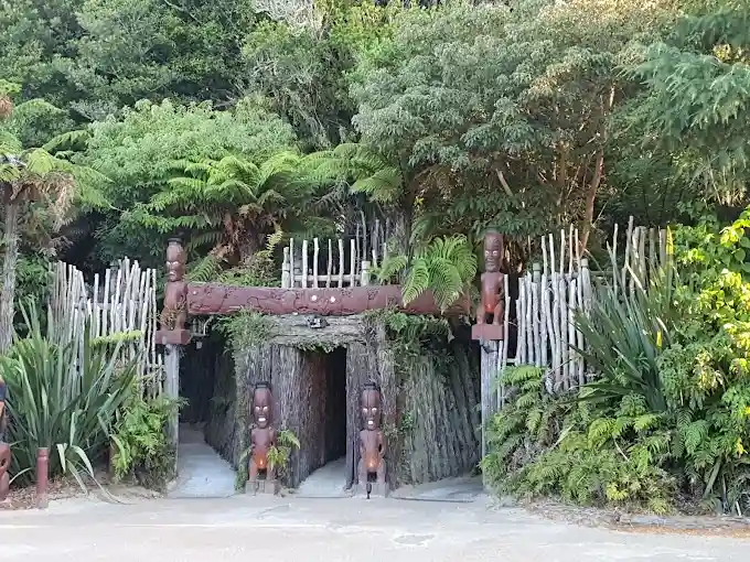a photograph of a traditional maori meeting house entrance, featuring carved wooden figures and a wooden gate, surrounded by…