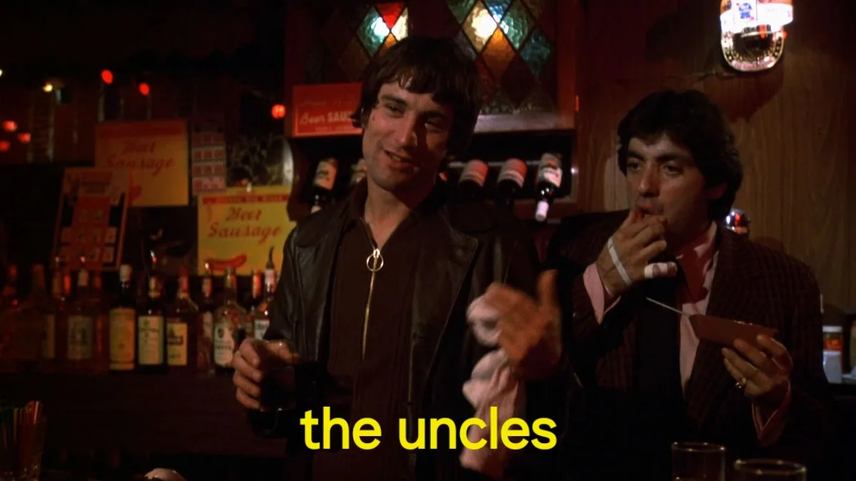 Two men sit at a dimly lit bar; the man on the left smiles while holding a drink, and the man on the right eats a sandwich…