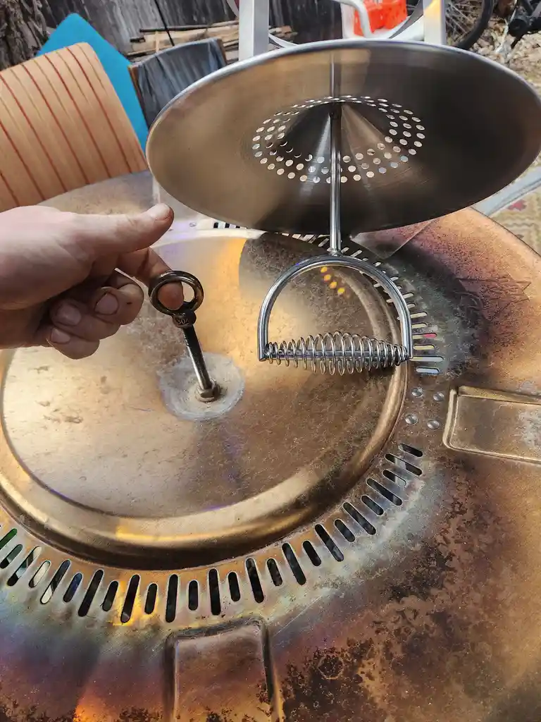 A hand holds a small key next to a metal grill lid with a spring-loaded latch mechanism, implying the grill was recently…
