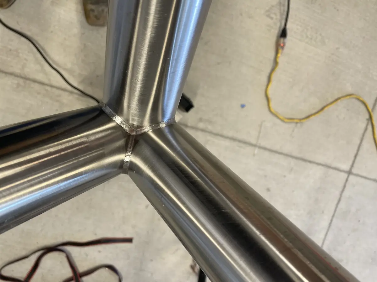 A close-up photo shows three brushed stainless steel pipes meeting at a central vertical pipe, with visible weld seams at…