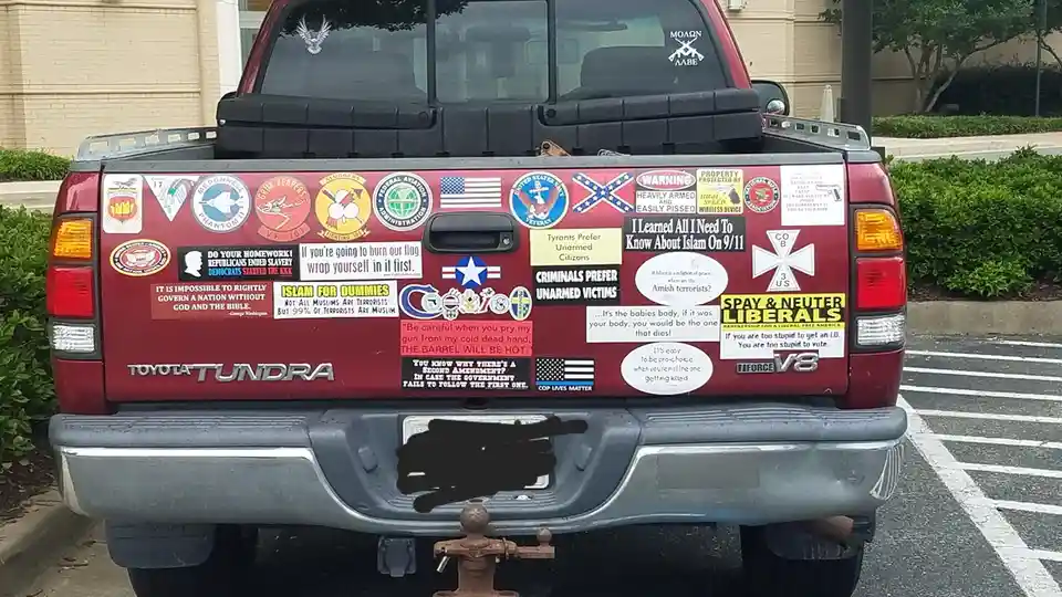 The rear of a red Toyota Tundra pickup truck is covered in numerous political and extremist stickers, including flags…