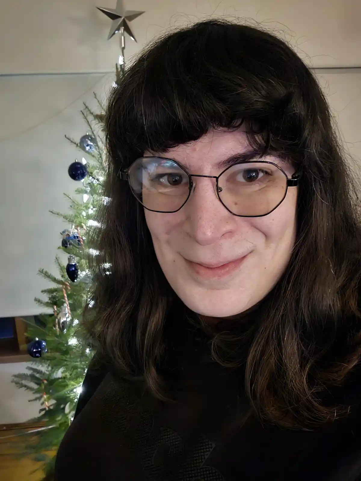 A person with dark hair and glasses smiles subtly while standing in front of a decorated Christmas tree with blue ornaments…
