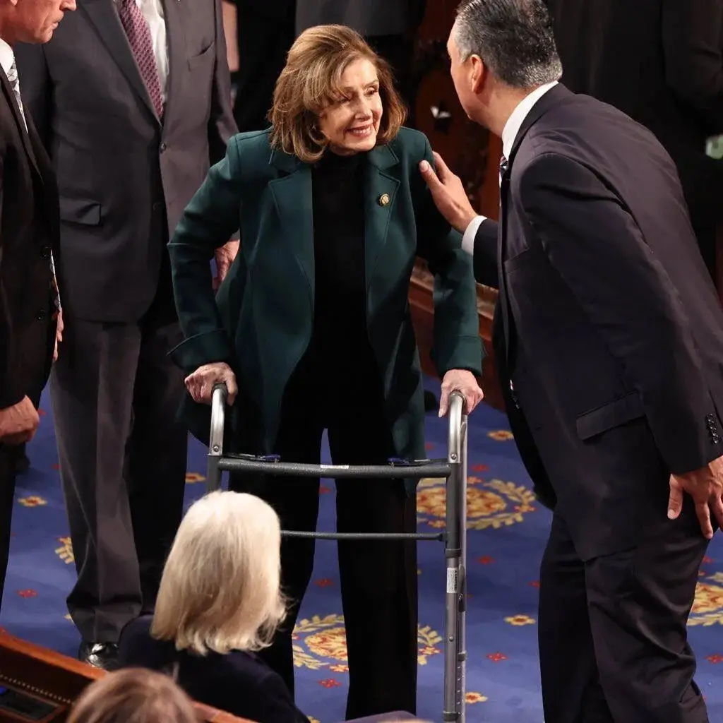 A woman in a dark green blazer and black pants uses a metal walker while smiling warmly as a man in a suit gently touches…