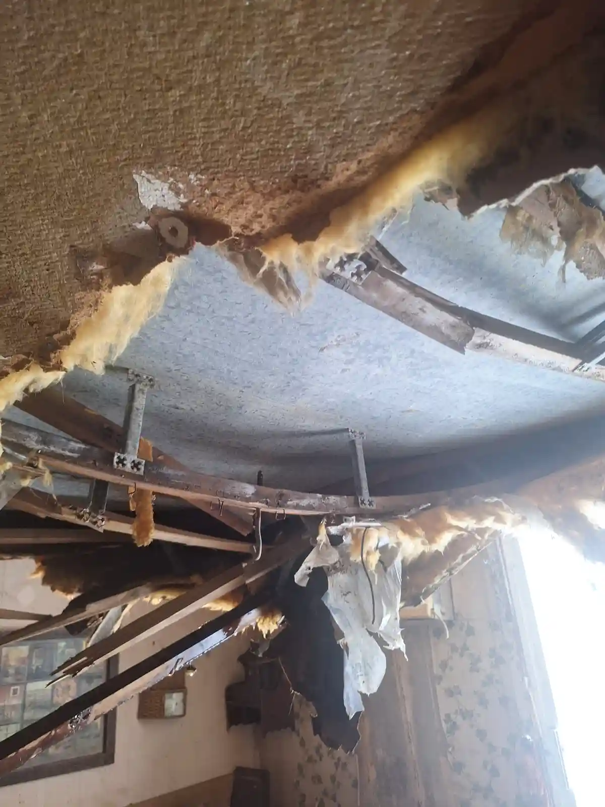 A severely damaged ceiling with large holes, exposed yellow insulation, and broken wooden beams hanging down. The overlaid…