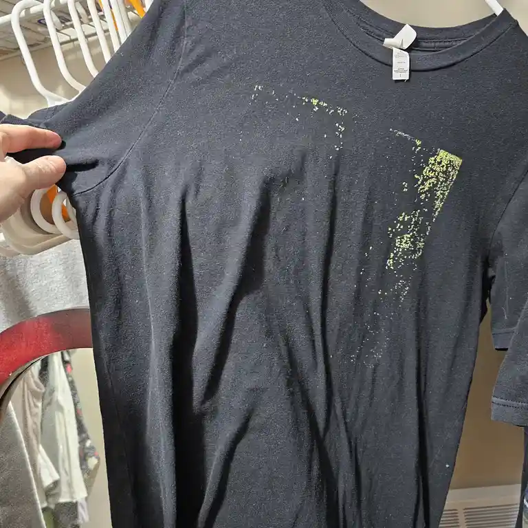 A person holds up a dark gray t-shirt on a white hanger, revealing a faded, glittery design that looks like a stylized '7'…