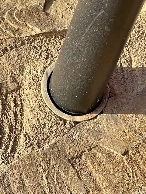 A close-up photo shows a dark, cylindrical metal pole set into a rough, textured stone or concrete surface. The pole is…