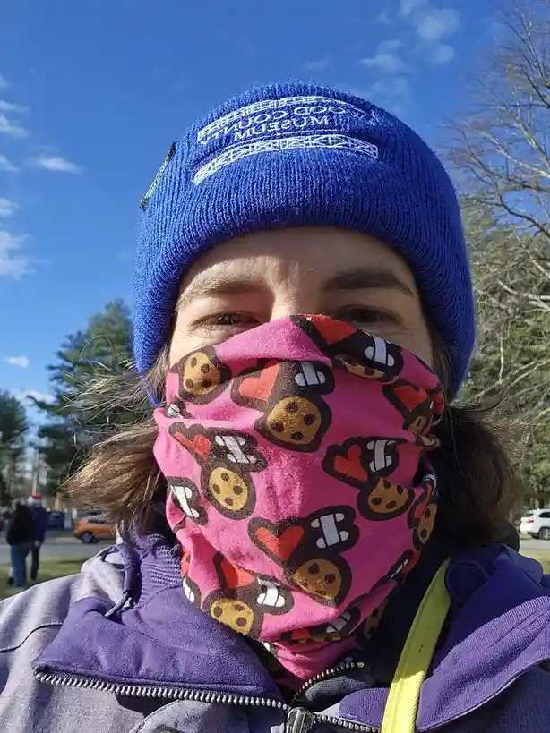 A person outdoors in bright sunlight wears a blue beanie with 'GOOD COUNTY MUSEUM' embroidered on it and a pink face…