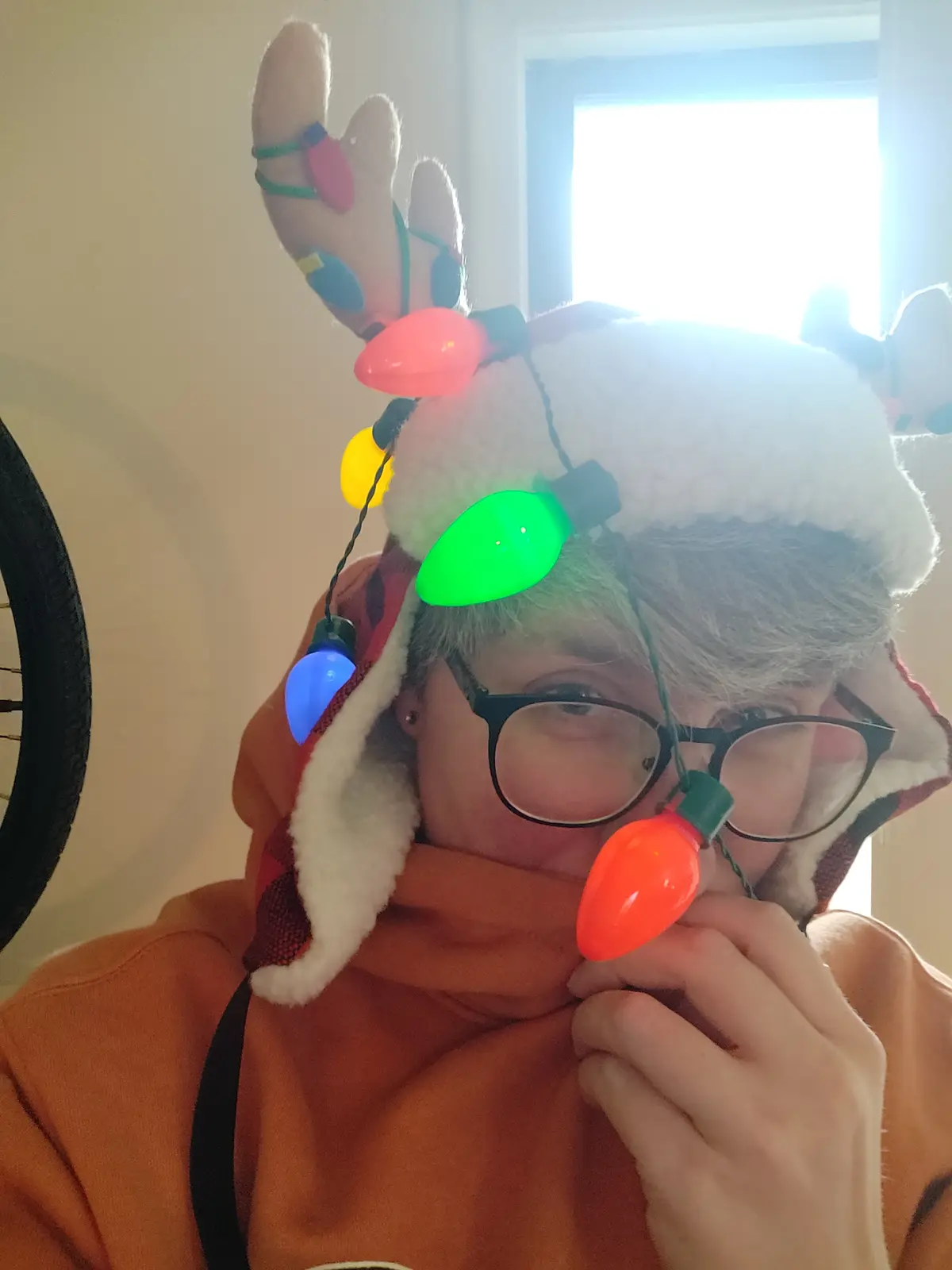 A person wearing a reindeer-themed hat with glowing, multi-colored Christmas lights tangled around their head and face…