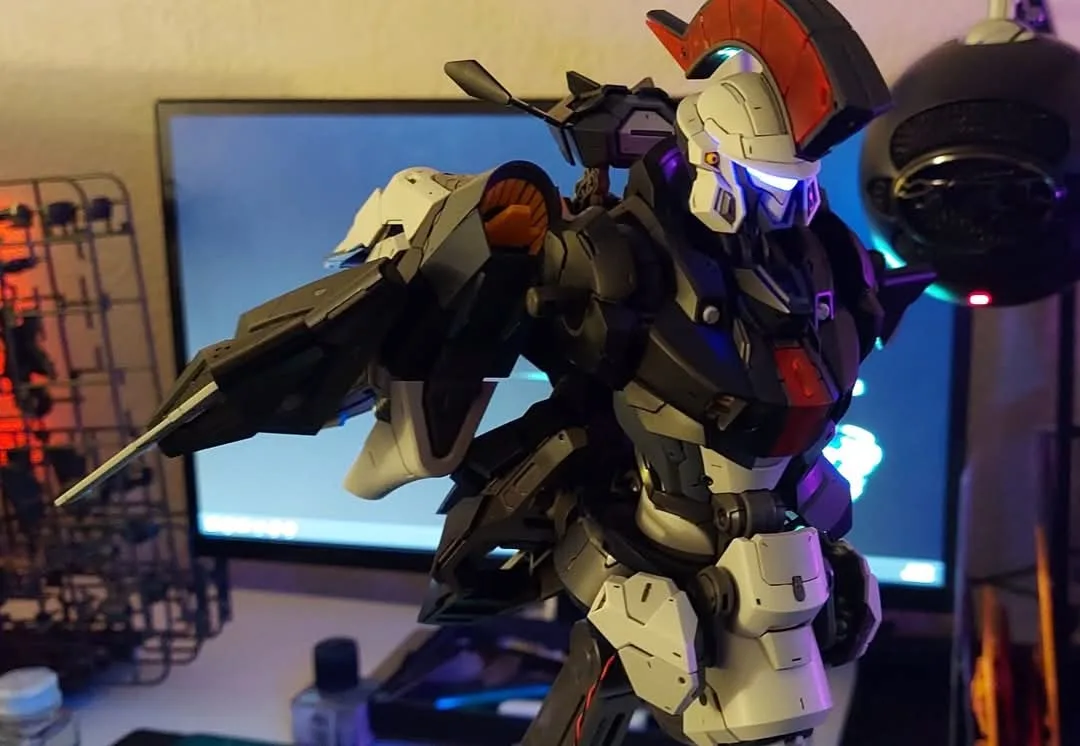A detailed, white, black, and red Gundam model stands in a dynamic pose on a desk, holding a weapon, with a glowing blue…