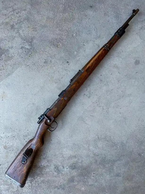 A wooden-stocked bolt-action rifle with a metal barrel and trigger mechanism lies diagonally on a gray concrete floor. The…