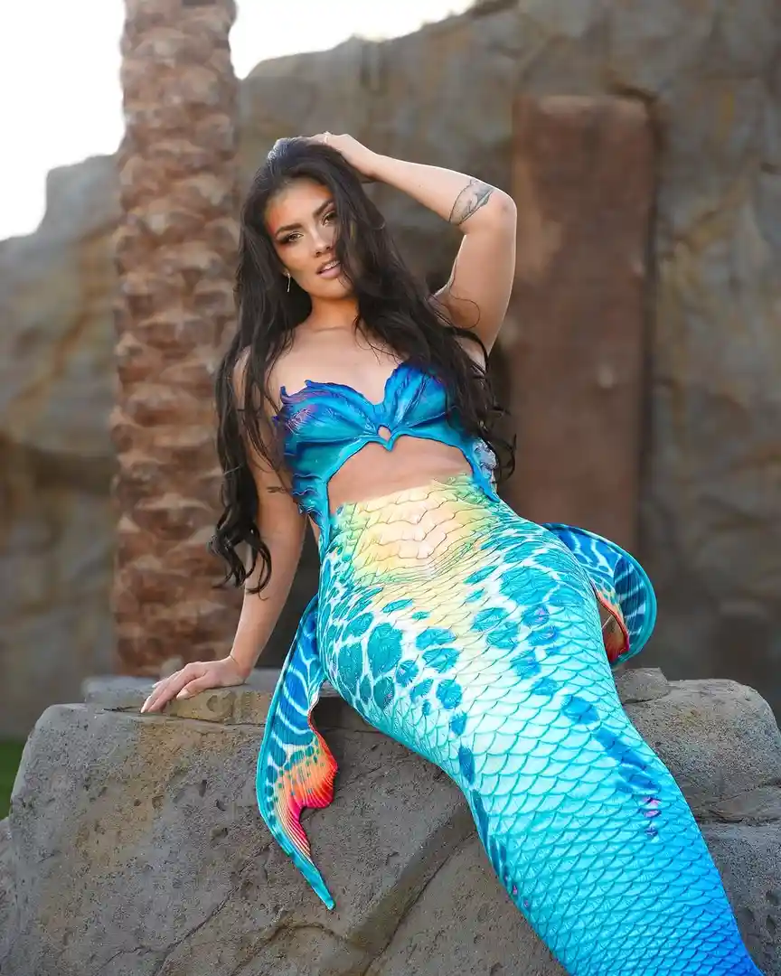 A woman with long dark hair is posing on a rock wearing a colorful mermaid tail costume with blue, green, and yellow scales…