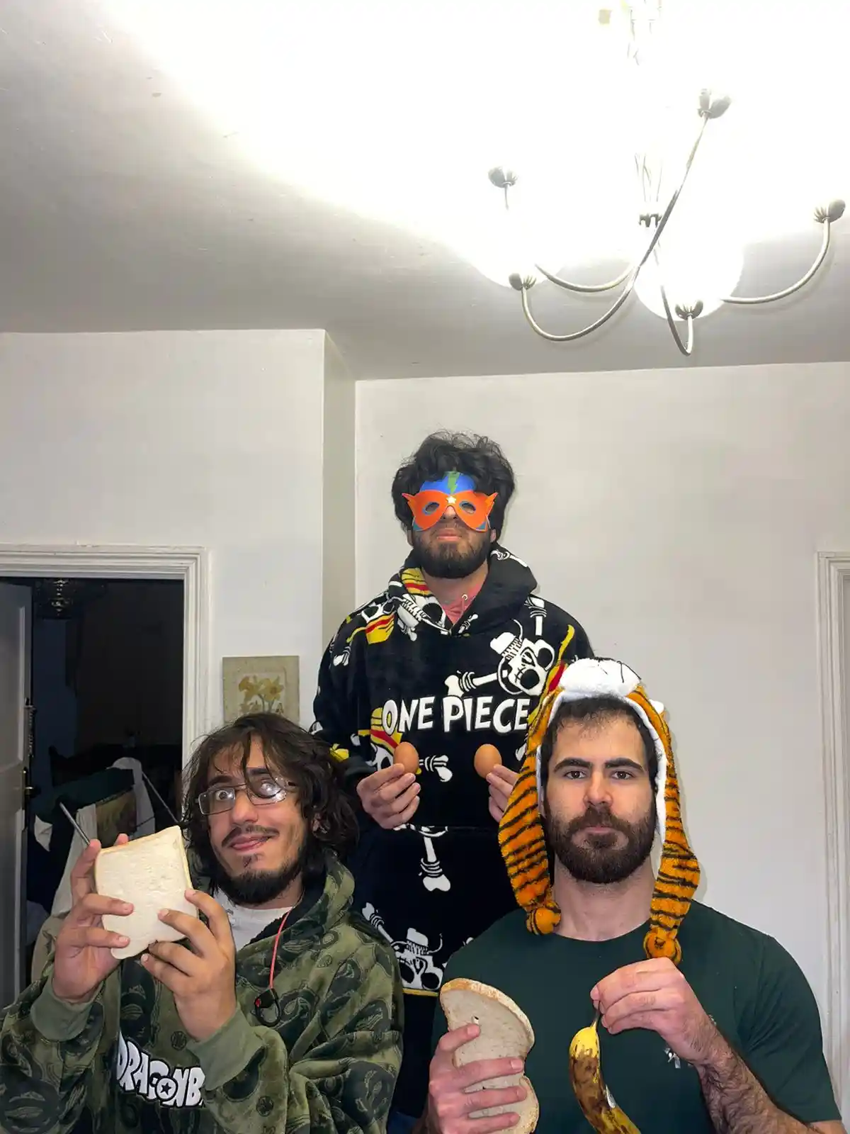 Three men pose indoors, one in a tiger hat holding bread and a banana, another in a green hoodie holding a slice of bread…