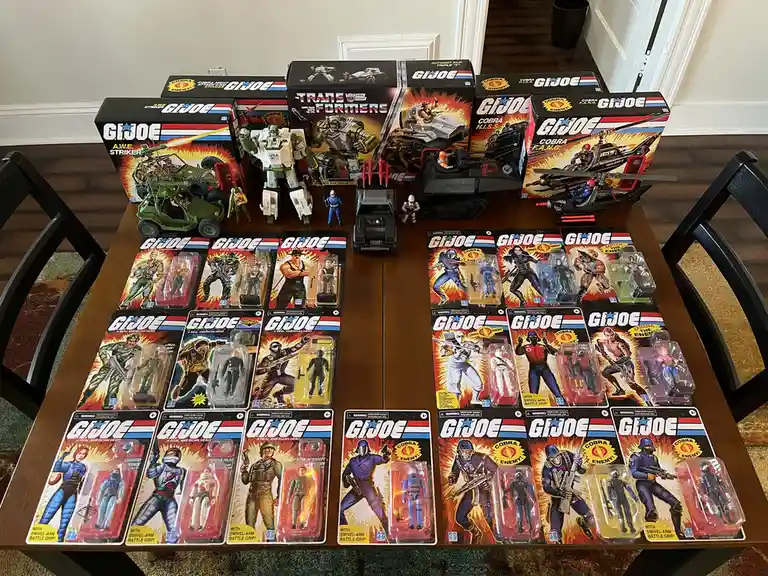 A wooden table is covered with numerous vintage G.I. Joe action figures in their original blister packs and several large…