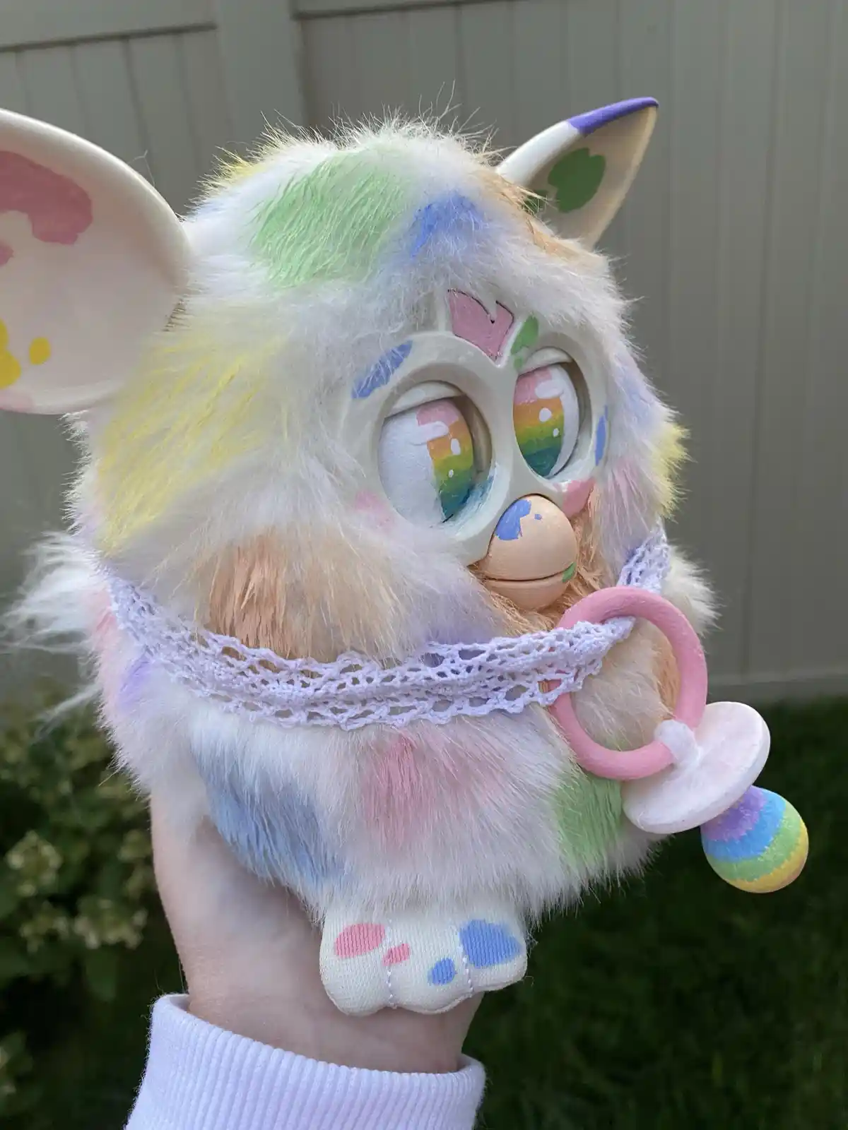 A hand holds a heavily customized, pastel-colored Furby toy with rainbow-painted fur, large rainbow-reflective eyes, and a…