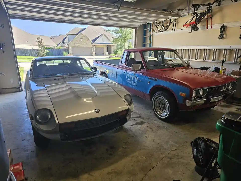 Two vintage cars are parked inside a residential garage: a silver Datsun 240Z on the left and a red-and-blue Datsun pickup…