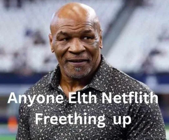 A close-up photo of Mike Tyson smiling slightly while looking off to the side, wearing a patterned shirt, with the text…