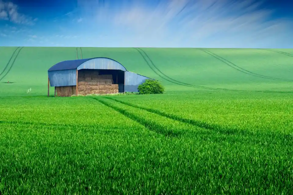 A serene rural landscape features a blue metal barn filled with hay bales, set against rolling green fields with visible…