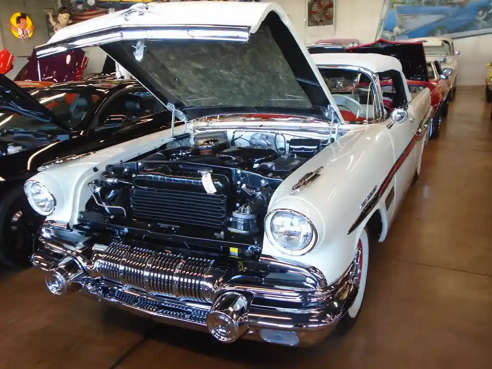 A white 1950s classic car with its hood open, revealing a shiny engine, is parked in a showroom with other vintage cars. The…