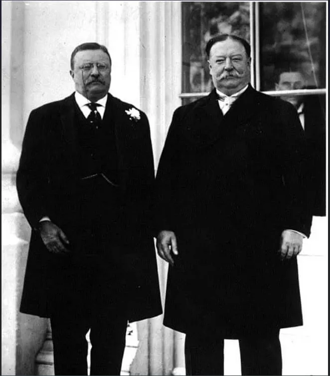 a black-and-white historical photograph shows two men in formal overcoats standing side by side outdoors. The man on the…