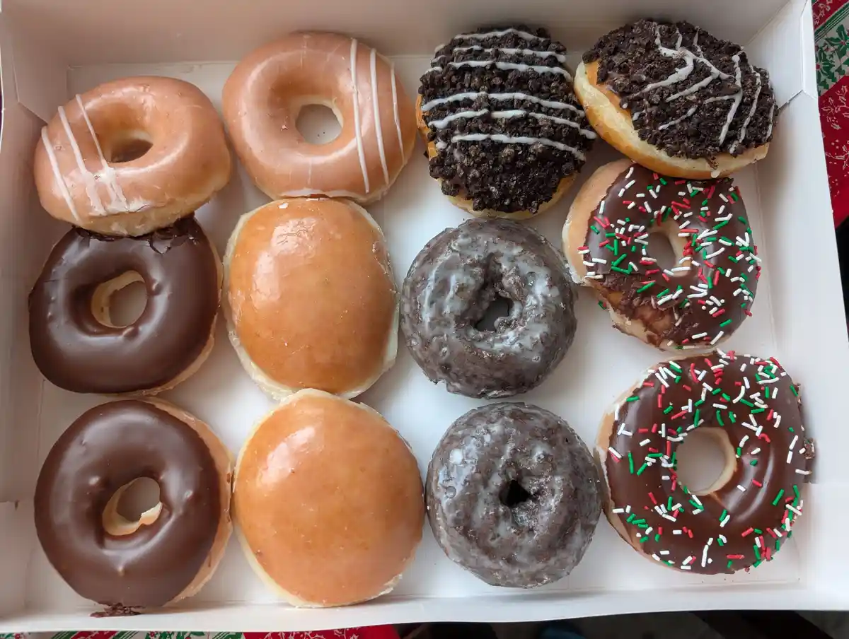 A top-down view of a white cardboard box filled with twelve assorted donuts, including glazed, chocolate frosted, chocolate…