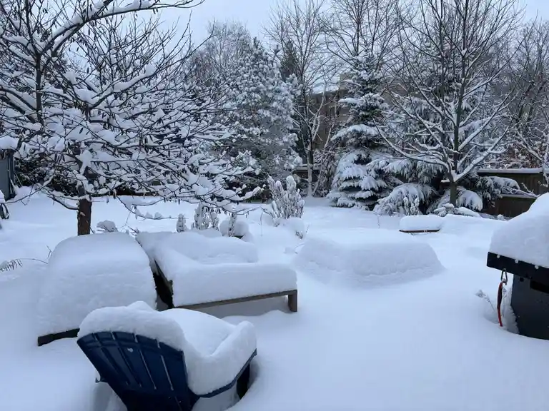 A backyard is completely buried under deep snow, with outdoor furniture like a blue Adirondack chair and wooden benches…