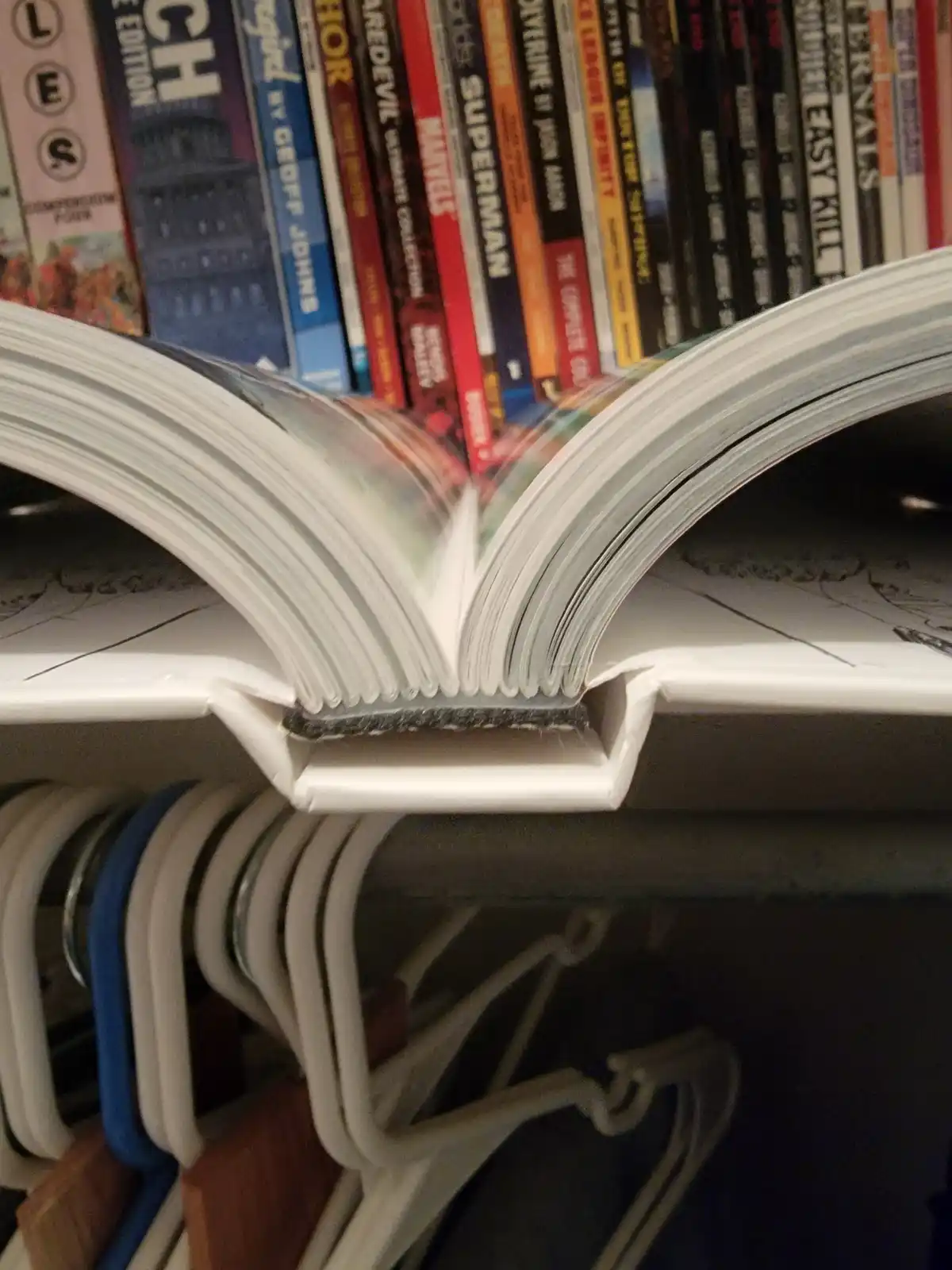 An open comic book rests on a white shelf, its pages fanned out, with a row of comic books visible behind it and clothes…
