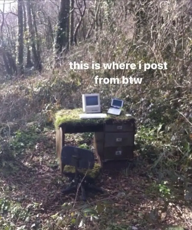 An old wooden desk covered in moss sits in a forest, with a vintage beige computer and a small laptop placed on top. A black…