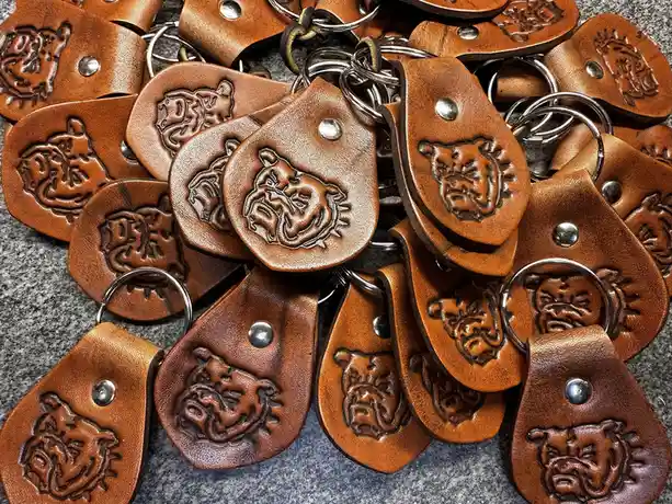A pile of handmade brown leather keychains, each featuring an embossed bulldog head design and a metal rivet, scattered on a…