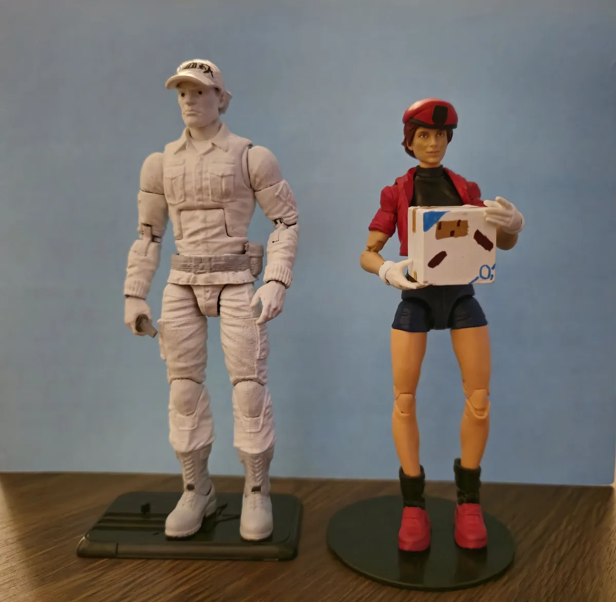 Two action figures stand side by side on a wooden surface against a blue wall. The figure on the left is entirely painted…