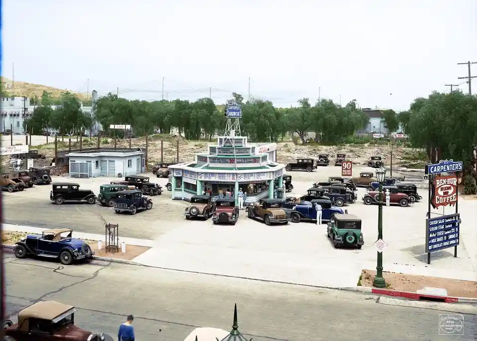 A vintage colorized photo shows a 1930s-era 'Carpenter's' drive-in restaurant with numerous classic cars parked around it…