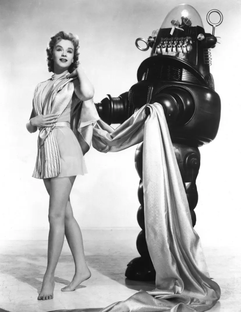 A vintage black-and-white photo shows a smiling woman in a short, shiny dress standing next to a large, retro-futuristic…